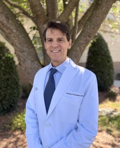 Portrait of Dr. Stephen A. Rendulich, DDS wearing a light blue lab coat and dark tie, posing outdoors in front of trees